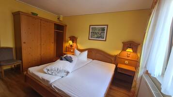 Habitación de hotel con una cama de madera, dos mesitas de noche con lámparas, un armario, una silla y un cuadro en la pared.