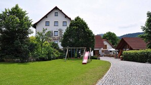 A building with a white facade and a red roof, surrounded by a well-maintained lawn and a cobblestone path. - Landhotel Zur Pferdetränke (Schleid)
