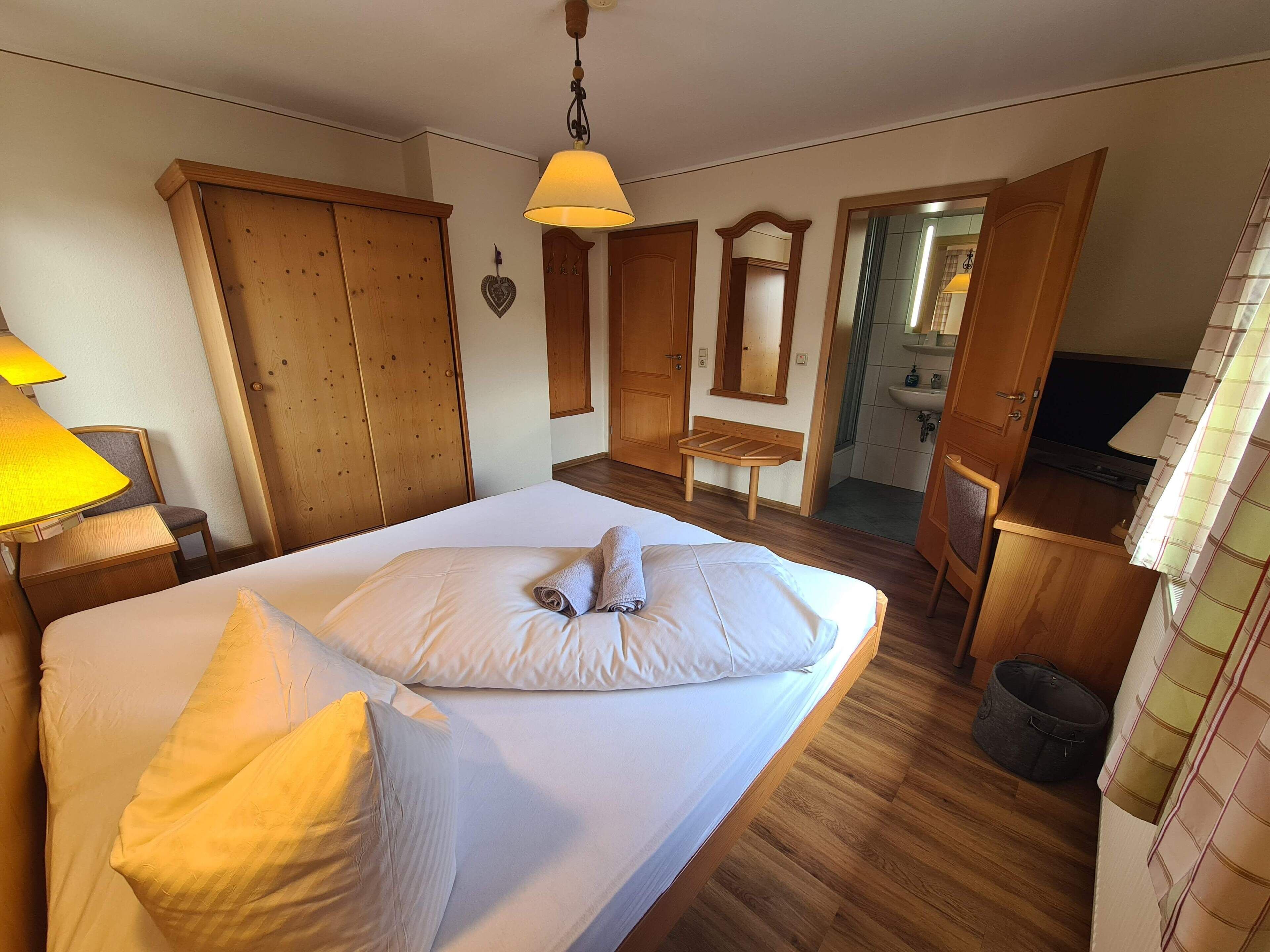 A bedroom with a bed, wooden wardrobe, a bench, a mirror, and a bathroom visible through an open door.