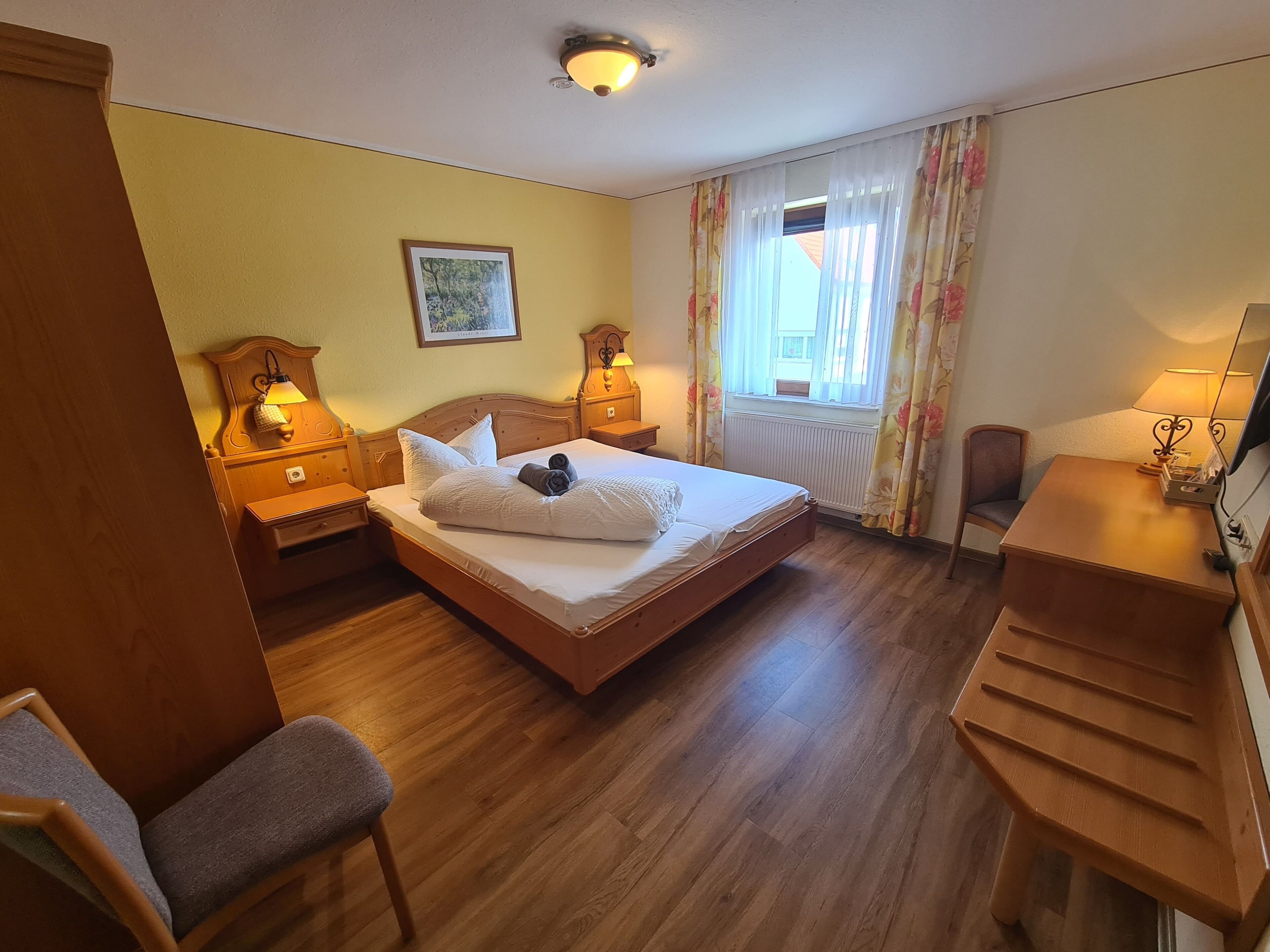 Habitación de hotel con una cama de madera, dos mesitas de noche con lámparas, un escritorio, una silla y una ventana con cortinas florales.