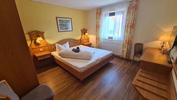Habitación de hotel con una cama de madera, dos mesitas de noche con lámparas, un escritorio, una silla y una ventana con cortinas florales.