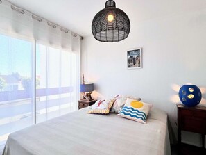 A bedroom with a bed, a hanging light fixture, a window with curtains, a wall-mounted picture, and a nightstand with a decorative object. - Charming Holiday Home with Garden View, Terrace, Balcony & Parking in Royan (Royan)