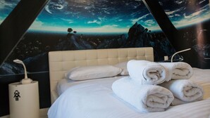 Un lit bien fait, avec des serviettes pliées, une tête de lit blanche et une fresque murale représentant un paysage montagneux sous un ciel étoilé.