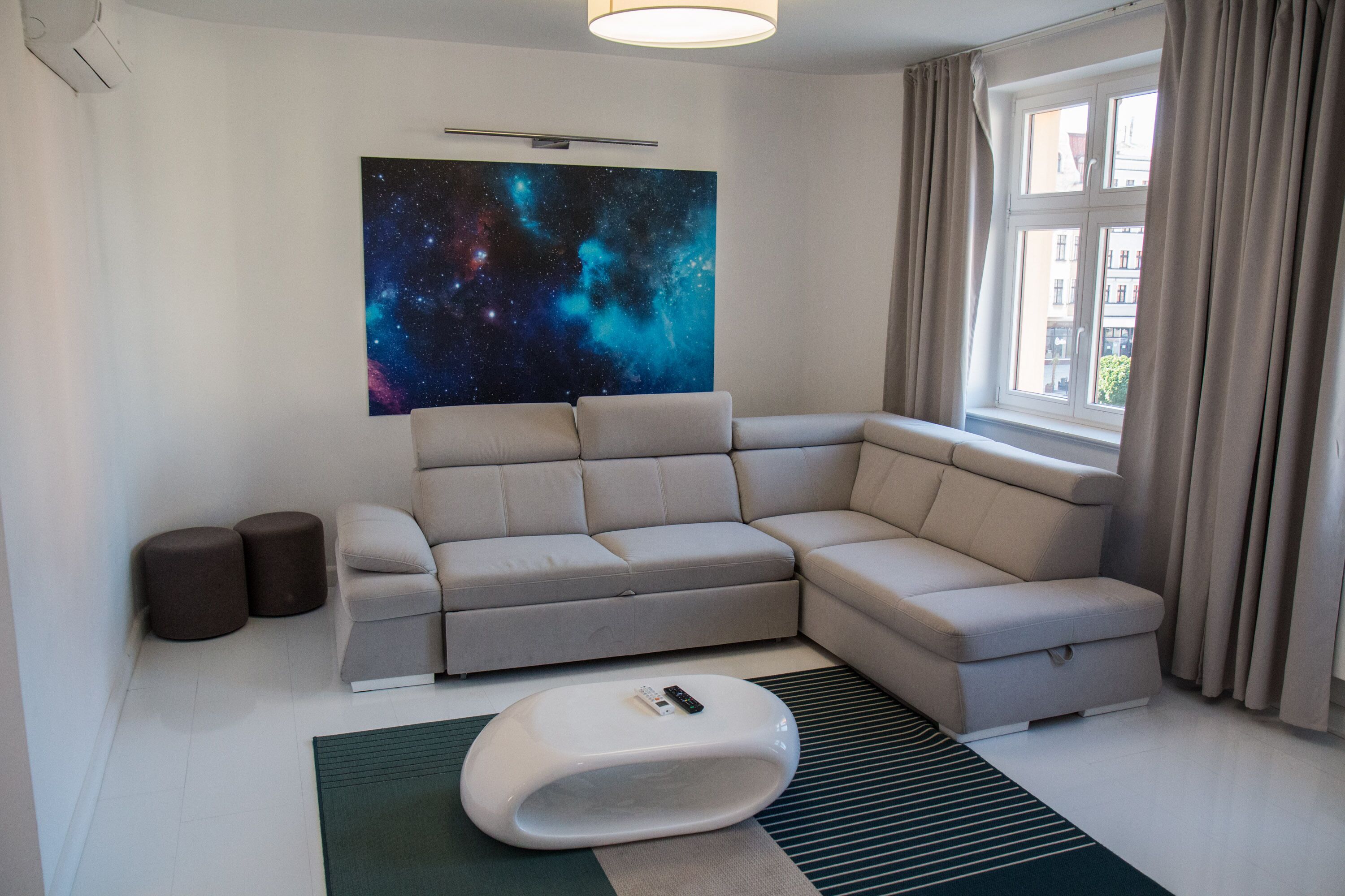 Een moderne woonkamer met een hoekbank, een unieke witte poef, wanddecoratie met een sterrenstelsel thema en een raam met gordijnen.