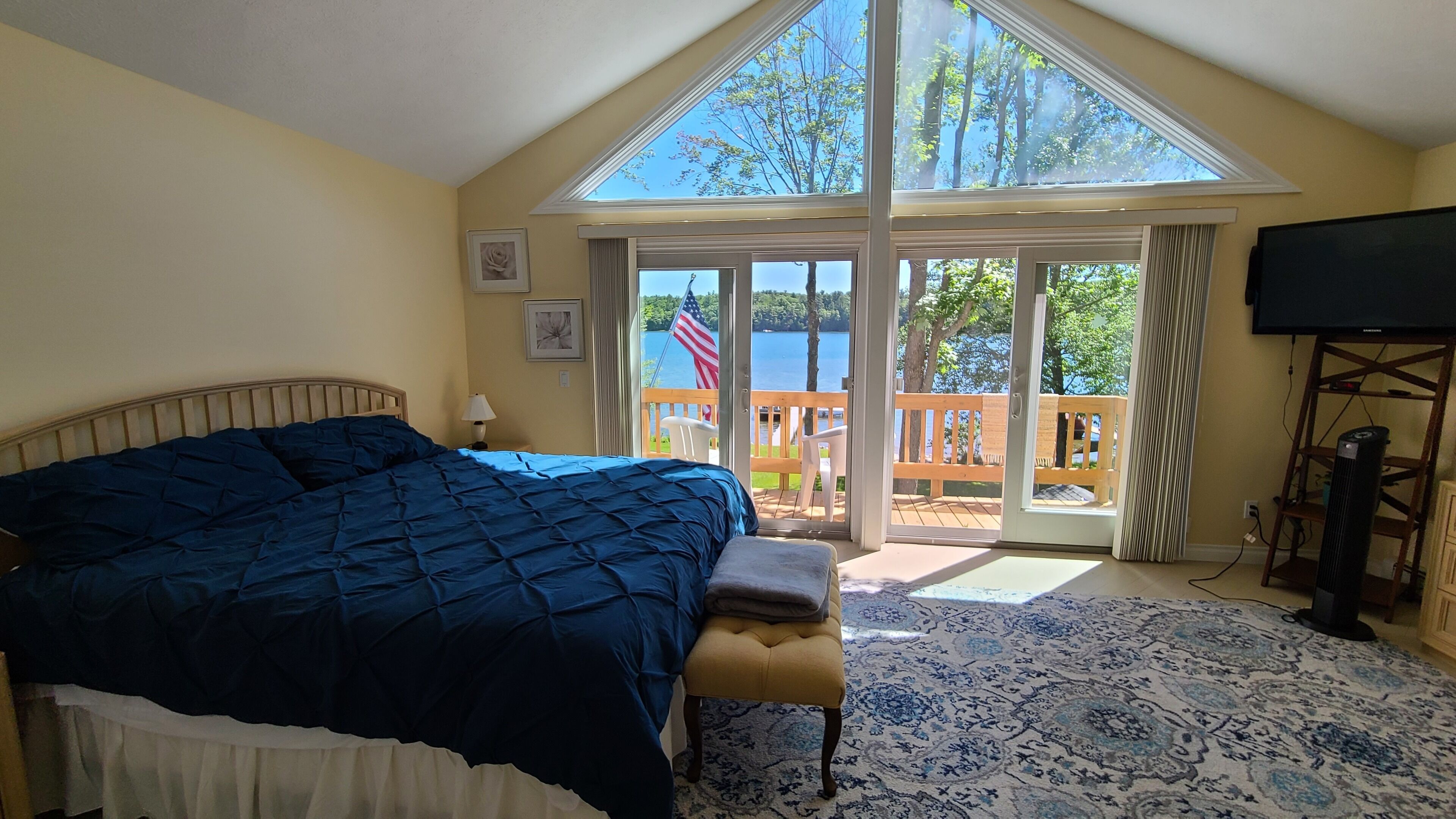 A bedroom with a large bed, a bench, a view of a lake through a sliding glass door, and a television on a stand.