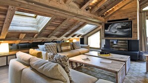 Un salon chaleureux de style loft, doté d’un haut plafond, de poutres apparentes, d’une grande fenêtre, d’un confortable ensemble de canapés, d’un téléviseur à écran plat et d’une cheminée.