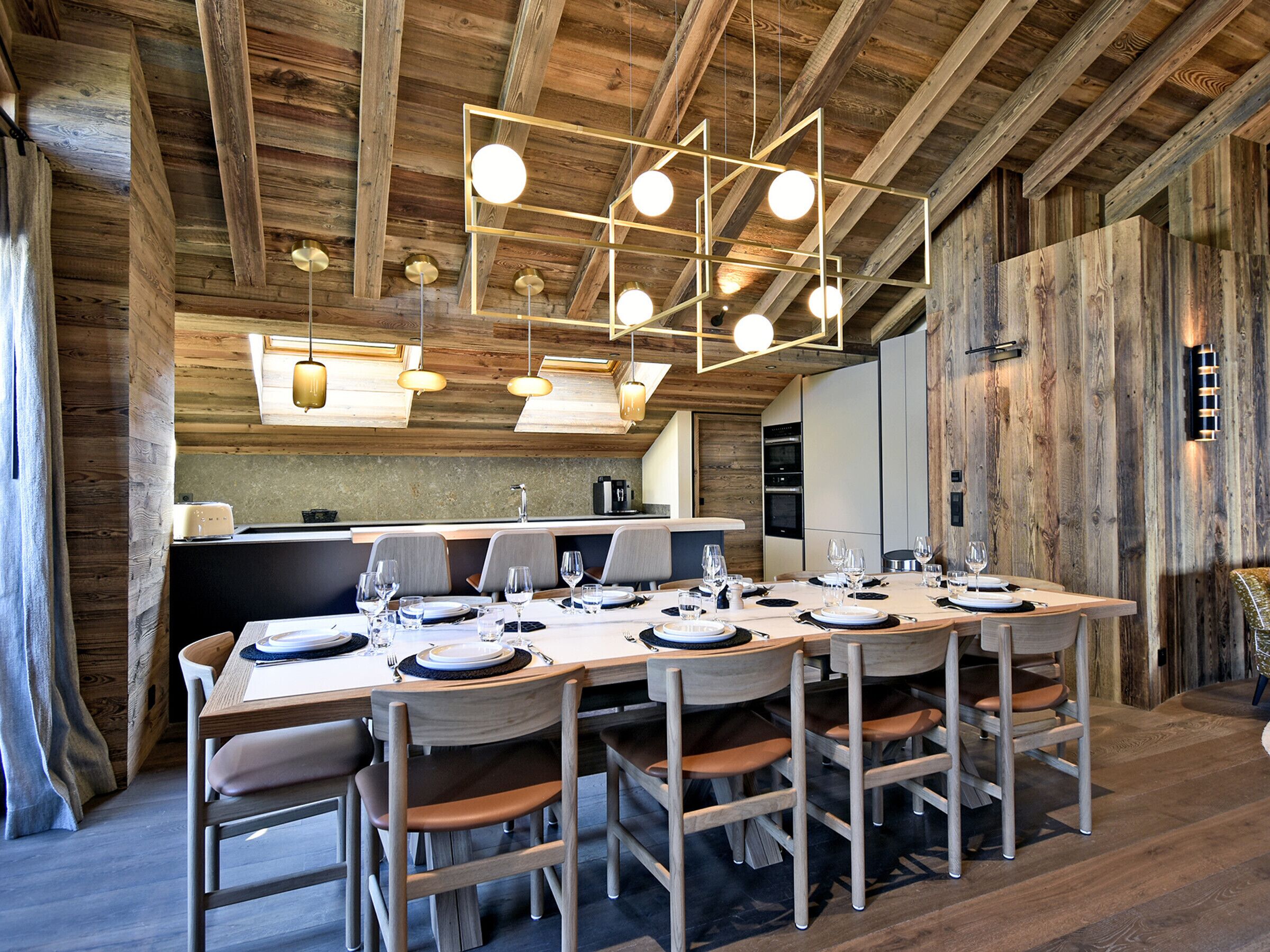 Un coin repas en bois avec une longue table dressée pour le repas, une cuisine en arrière-plan et un plafond rustique avec des poutres apparentes.