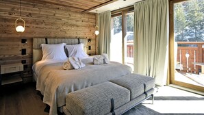 Une chambre avec un plafond en bois, un grand lit doté d’une tête de lit rembourrée de couleur grise, un banc et une porte-fenêtre coulissante donnant sur une terrasse extérieure.