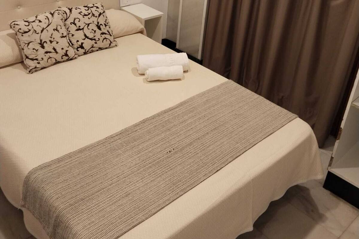 Un lit bien fait, recouvert d’une couvre-lit beige et agrémenté d’un coussin décoratif, une petite table de chevet avec une lampe, et un rideau qui recouvre une fenêtre.