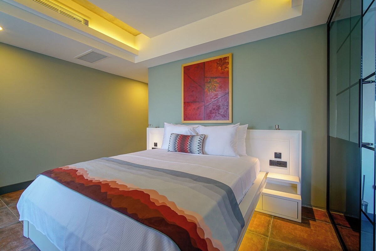 Habitación de hotel con una cama grande, una mesita de noche, una obra de arte colgada en la pared y un panel de vidrio.