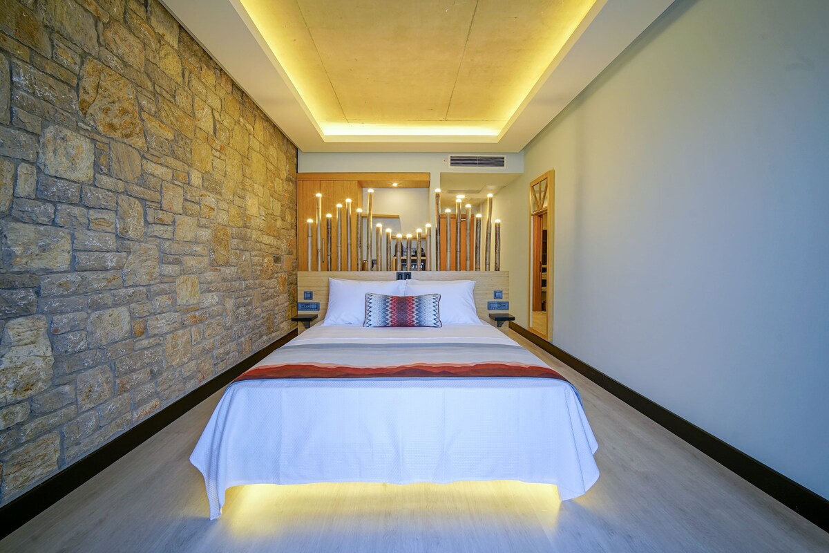 A modern hotel room with a stone wall, a large bed with a white bedspread, and a ceiling with recessed lighting.