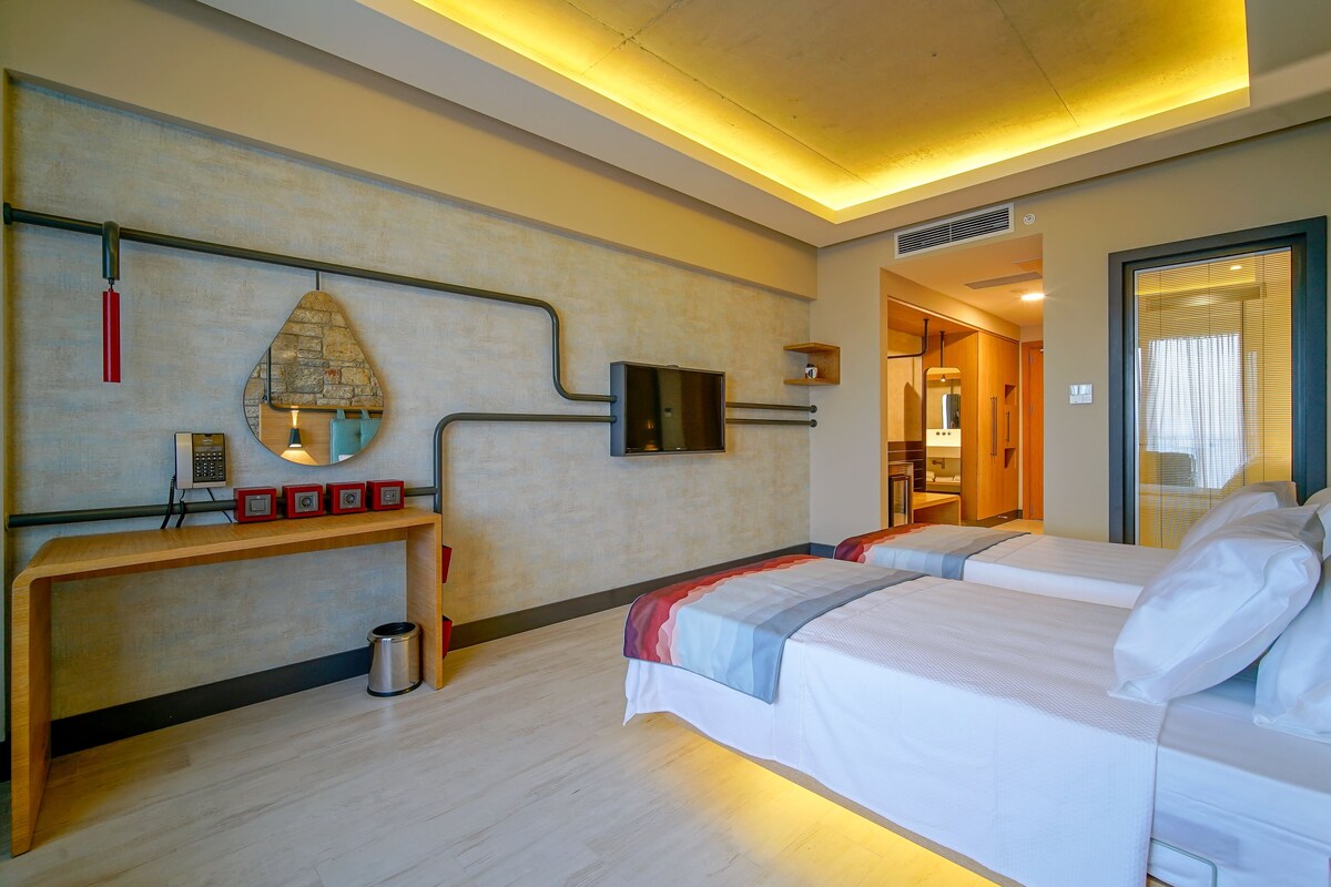 A hotel room with two beds, a wooden console table, a mirror, a television, and a window with blinds.