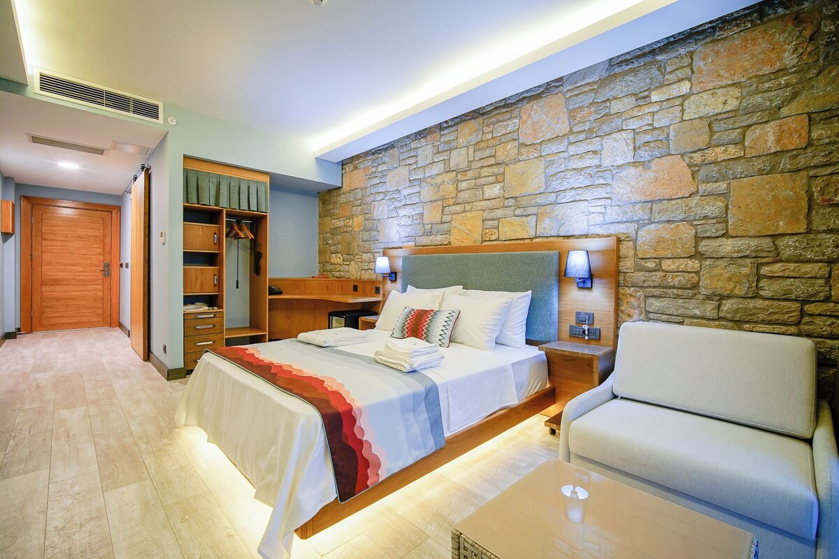 Habitación de hotel con pared de piedra, cama con cabecero, mesita de noche, una silla, armario y escritorio.