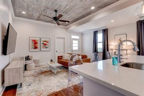 A modern kitchen with a central island, a dining area with a sofa, and a living room with a TV and artwork. - Updated 3BR Mid-City home with private pool. Steps to streetcar, near FQ & parade route. (New Orleans)