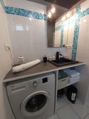 A compact hotel bathroom with a washing machine, a sink, and a mirror.