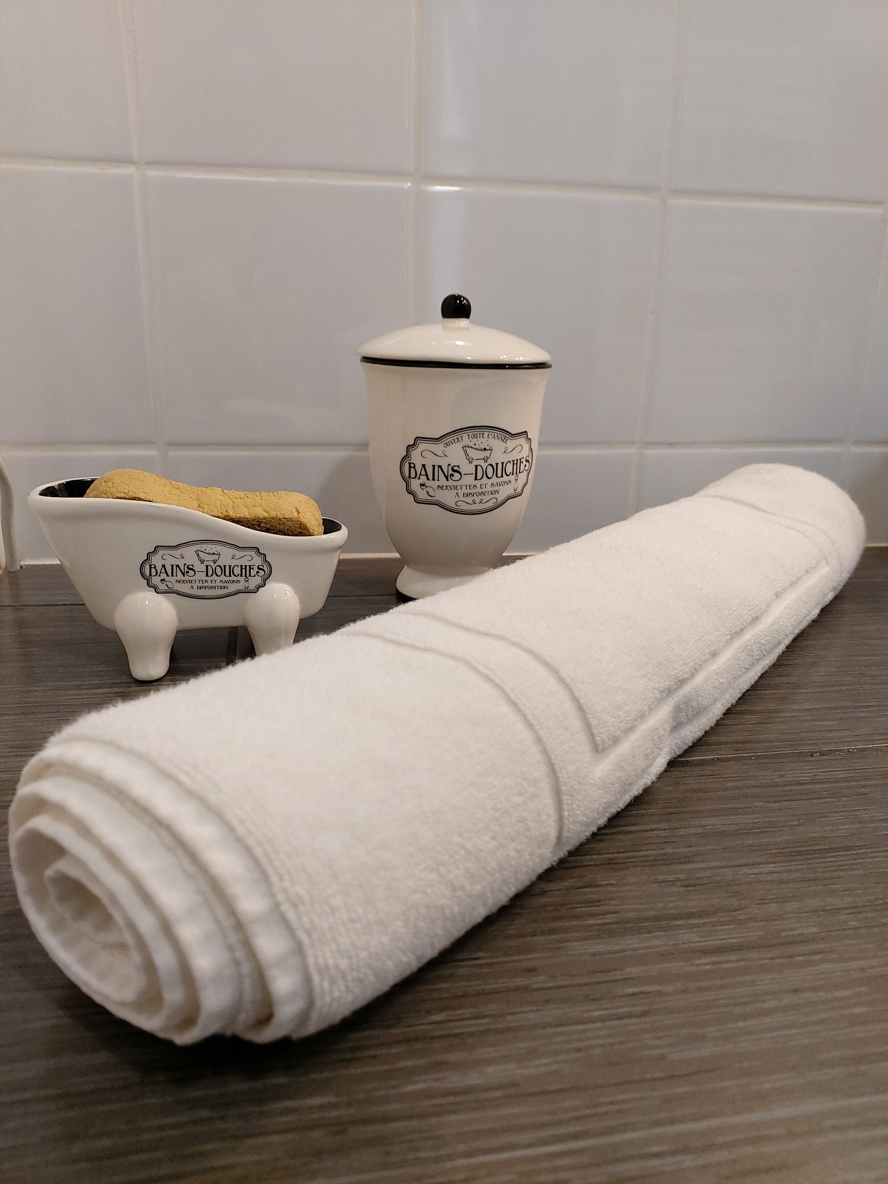 A white towel rolled up on a wooden surface, a small white ceramic container with a lid, and a smaller ceramic container with a lid.