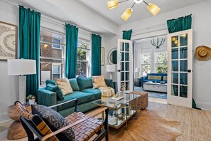 Un salon moderne avec un canapé vert, un fauteuil en cuir, une table basse en verre et une grande fenêtre ornée de rideaux turquoise.