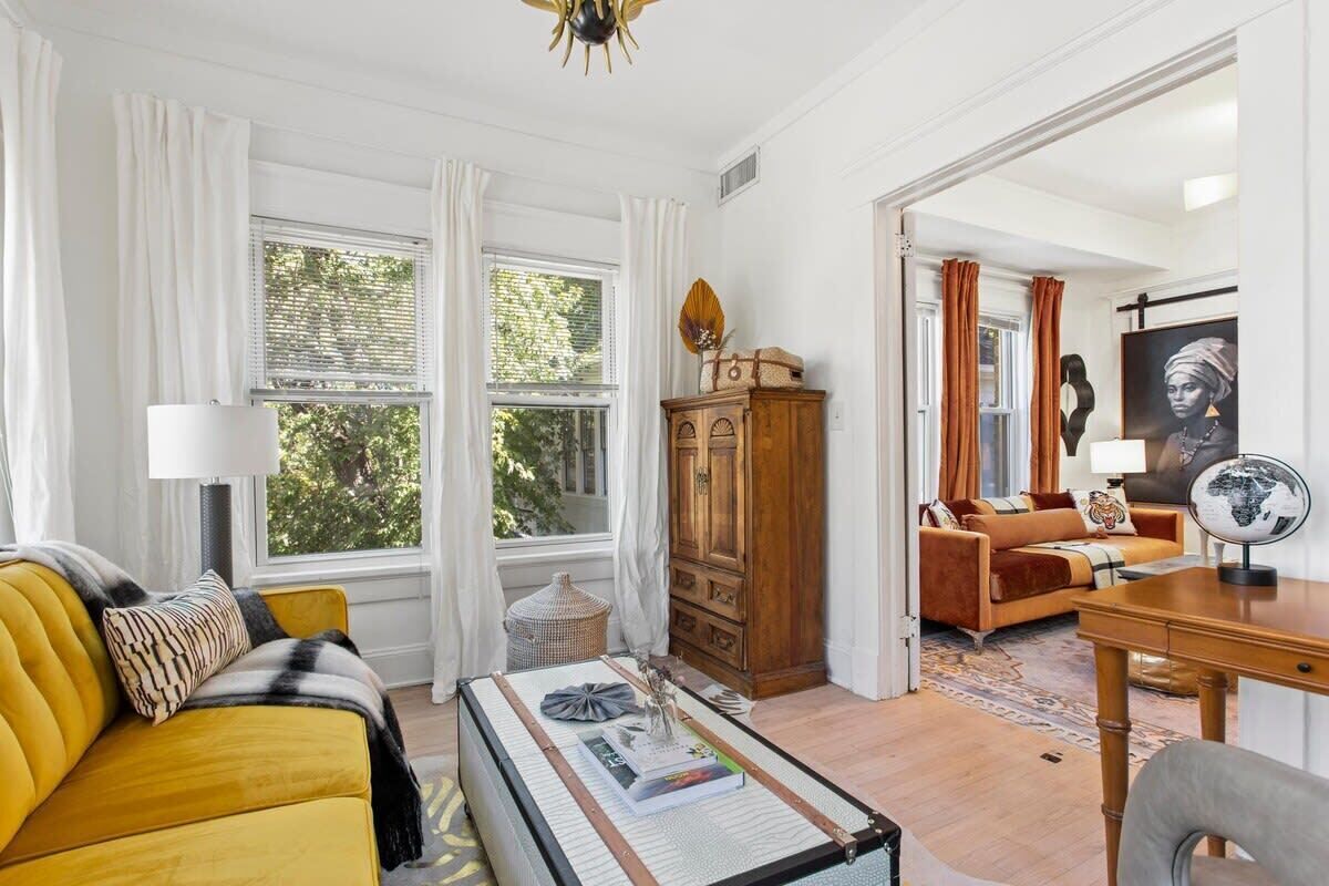 Una habitación con un sofá amarillo, un aparador de madera, un globo terráqueo sobre un escritorio y vistas al exterior a través de amplias ventanas.