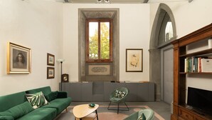 A room with a green sofa, a small table, a green chair, a bookshelf, and a large window with a view of trees. - Fantastic 3 bedroom apartment in the heart of Florence (Firenze)