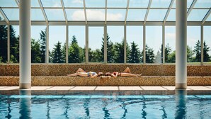 A swimming pool with a tiled edge, a mosaic-tiled bench, and a view of trees through large windows. - Pohorje Village Wellbeing Resort – Family Apartments Bolfenk (Maribor)