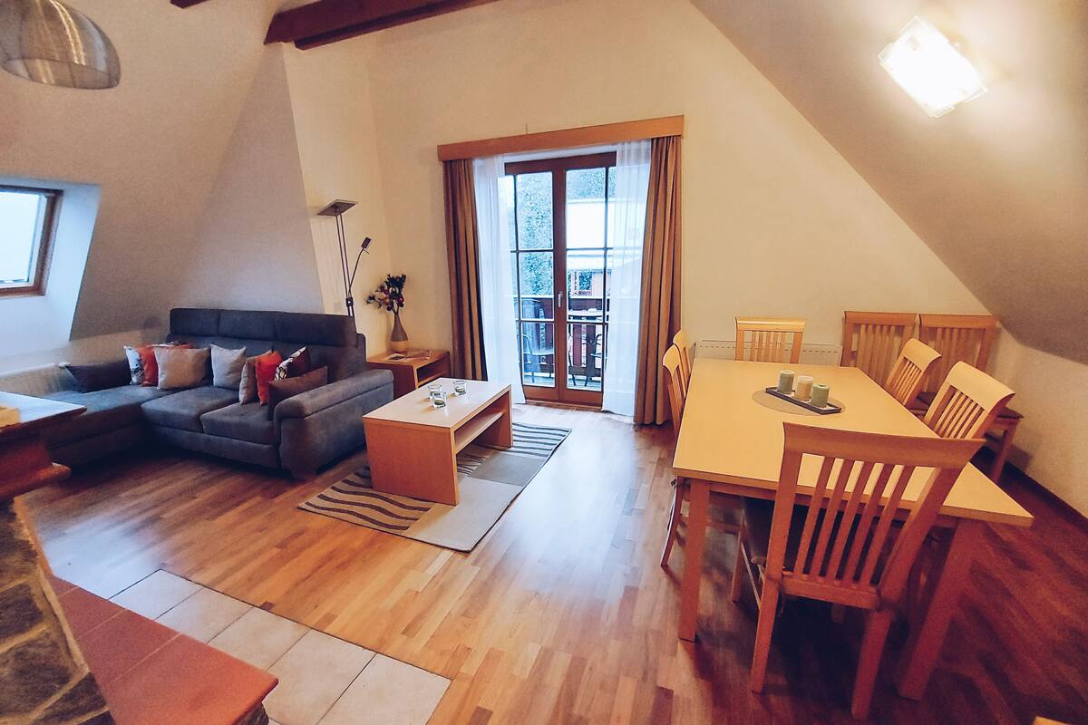Ett modernt vardagsrum med en soffa, ett soffbord, ett matbord och stolar. Det finns ett stort fönster med gardiner, en lampa och en vas med blommor på ett sidobord.