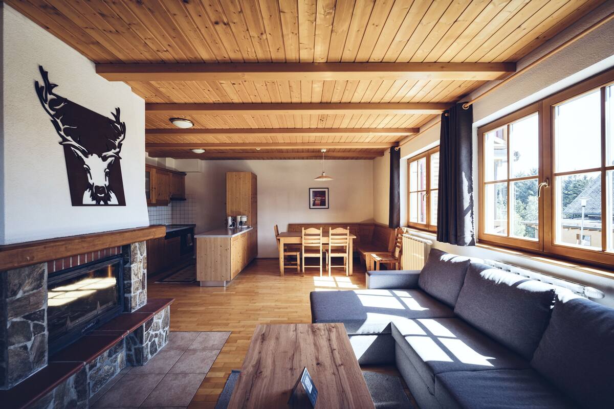 Ett mysigt vardagsrum med en steneldstad, träpanel i taket och stora fönster.