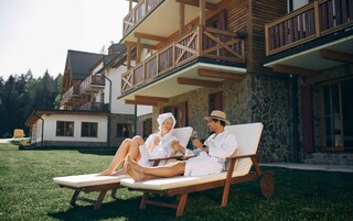 Una pareja descansando en una silla de madera en un césped bien cuidado, con un edificio de piedra y balcones al fondo.