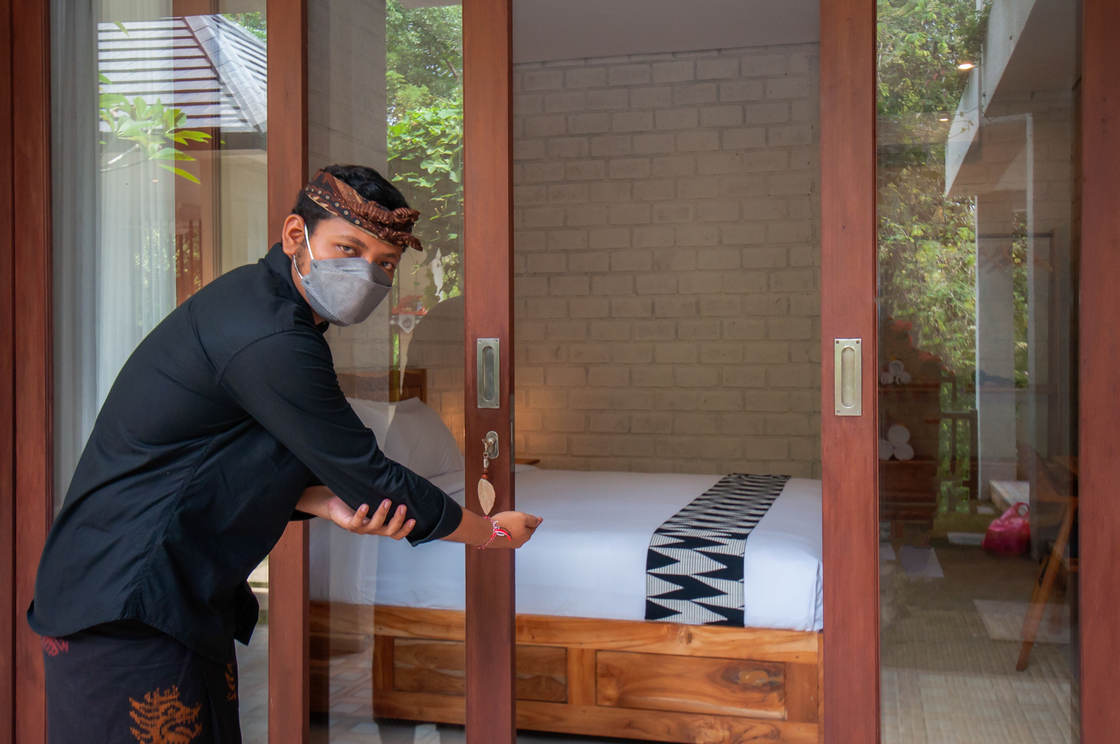 Una persona abre una puerta corrediza a un cuarto con una cama, un muro de ladrillo y vistas al exterior con vegetación.