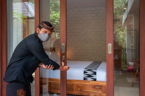 A person opening a sliding glass door to a room with a bed, a brick wall, and a view of greenery outside. - The Lokal Penida (Penida Island)