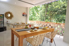 A dining area with a wooden table, wicker chairs, and a view of trees through large windows. - The Lokal Penida (Penida Island)