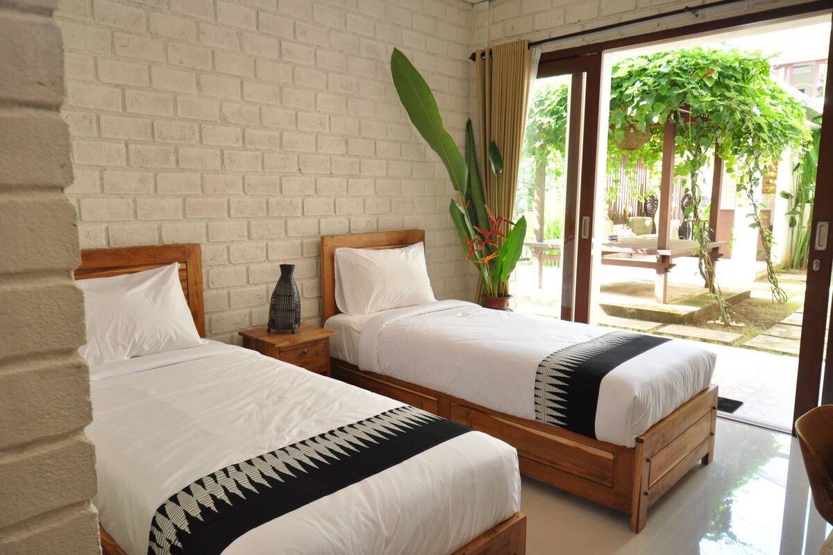 A room with two wooden beds, white bedding, a brick wall, a large potted plant, and a view of an outdoor garden.