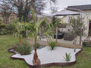 A patio area with a wooden deck, outdoor furniture, and a variety of plants including a palm tree and other greenery. - Pleasant house with spa (Périgueux)