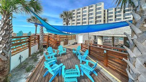 Una terraza de madera con sillas y toldo azules, rodeada de palmeras y un edificio con varias ventanas.