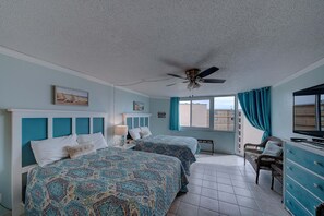 Ein Schlafzimmer mit zwei Betten, einem Deckenventilator, einem Fenster mit Vorhängen, einem Fernseher und einem Sessel.