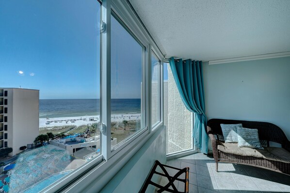 A balcony with a view of the beach and ocean, a wicker sofa, and a pool area.