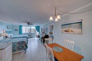 A room with two beds, a dining table, a ceiling fan, and a painting of a beach scene. - FREE Golf+Perks!Top of the Gulf Beachfront 703 Spacious Studio w/Great Views! (Panama City Beach)