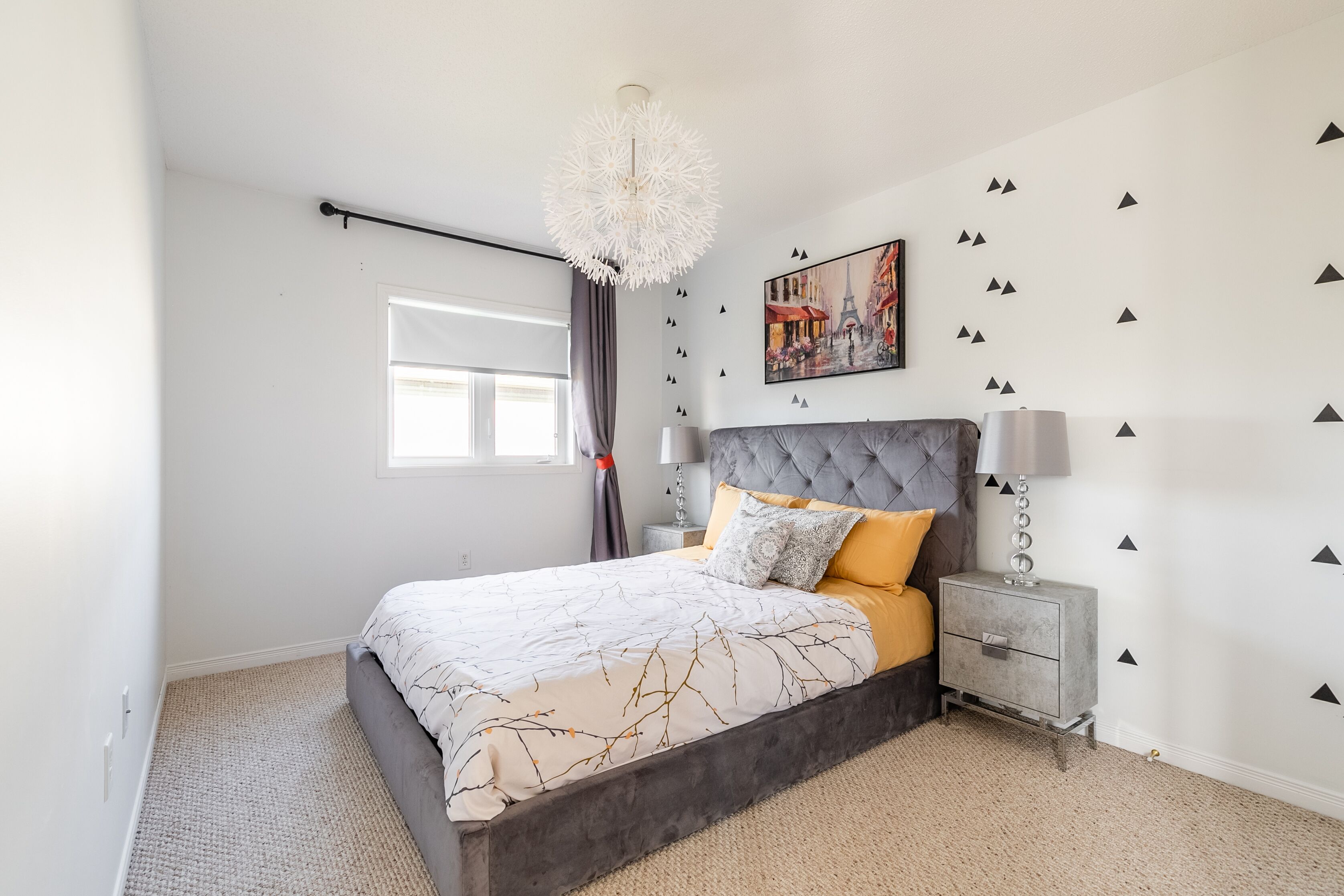 A bedroom with a bed, nightstands, a chandelier, a window with curtains, and wall decorations.