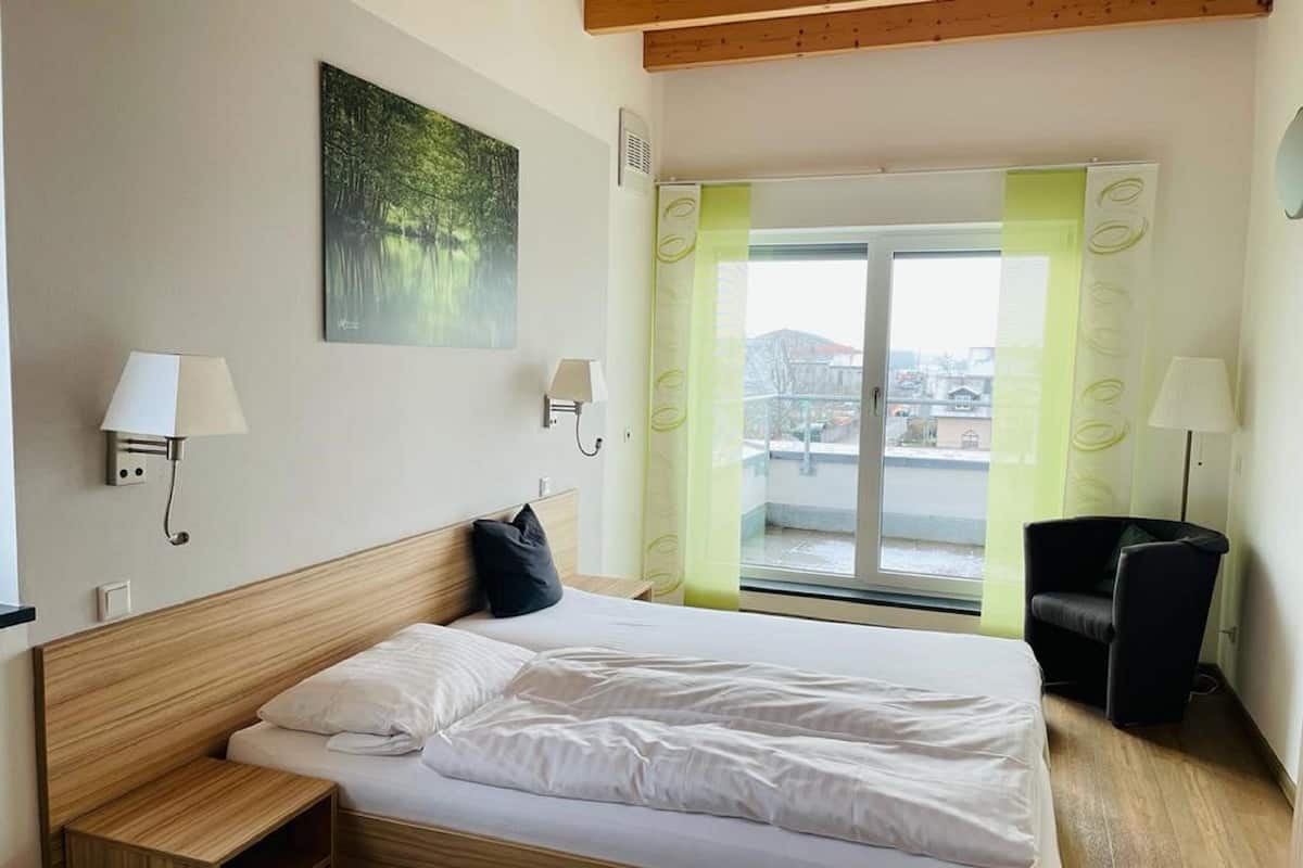A hotel room with a wooden bed, a nightstand, a black chair, a window with a view of buildings, and a green curtain.