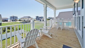 Una terraza techada con barandas blancas, una piscina y casas de playa al fondo.