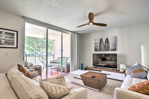 A modern living room with a sofa, coffee table, and TV. There is a balcony with chairs and a view of greenery. - Jupiter Bay Condo w/ Pool < Half Mile to Beach! (Jupiter)