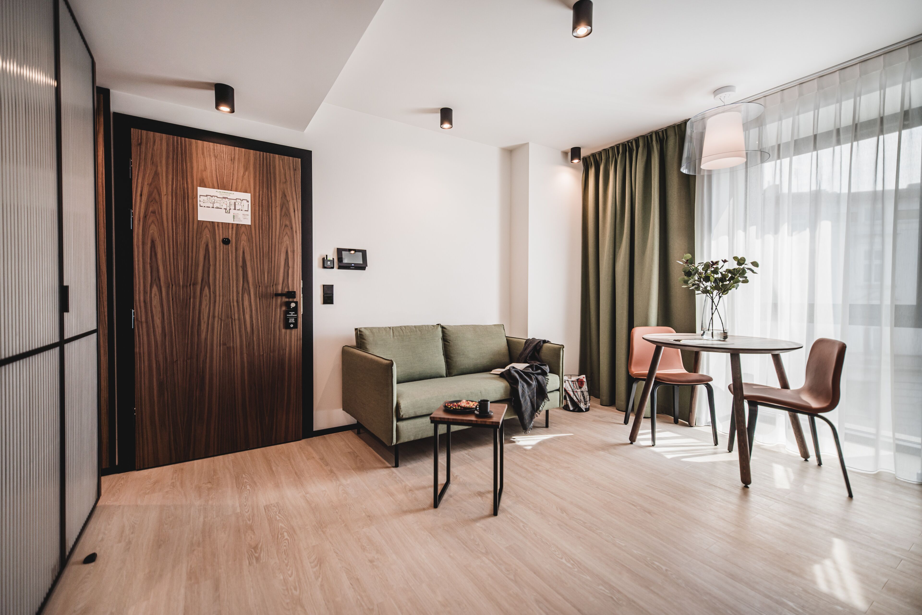 Una habitación de hotel moderna con una puerta de madera, un sofá verde, una mesita pequeña y una mesa redonda con sillas.