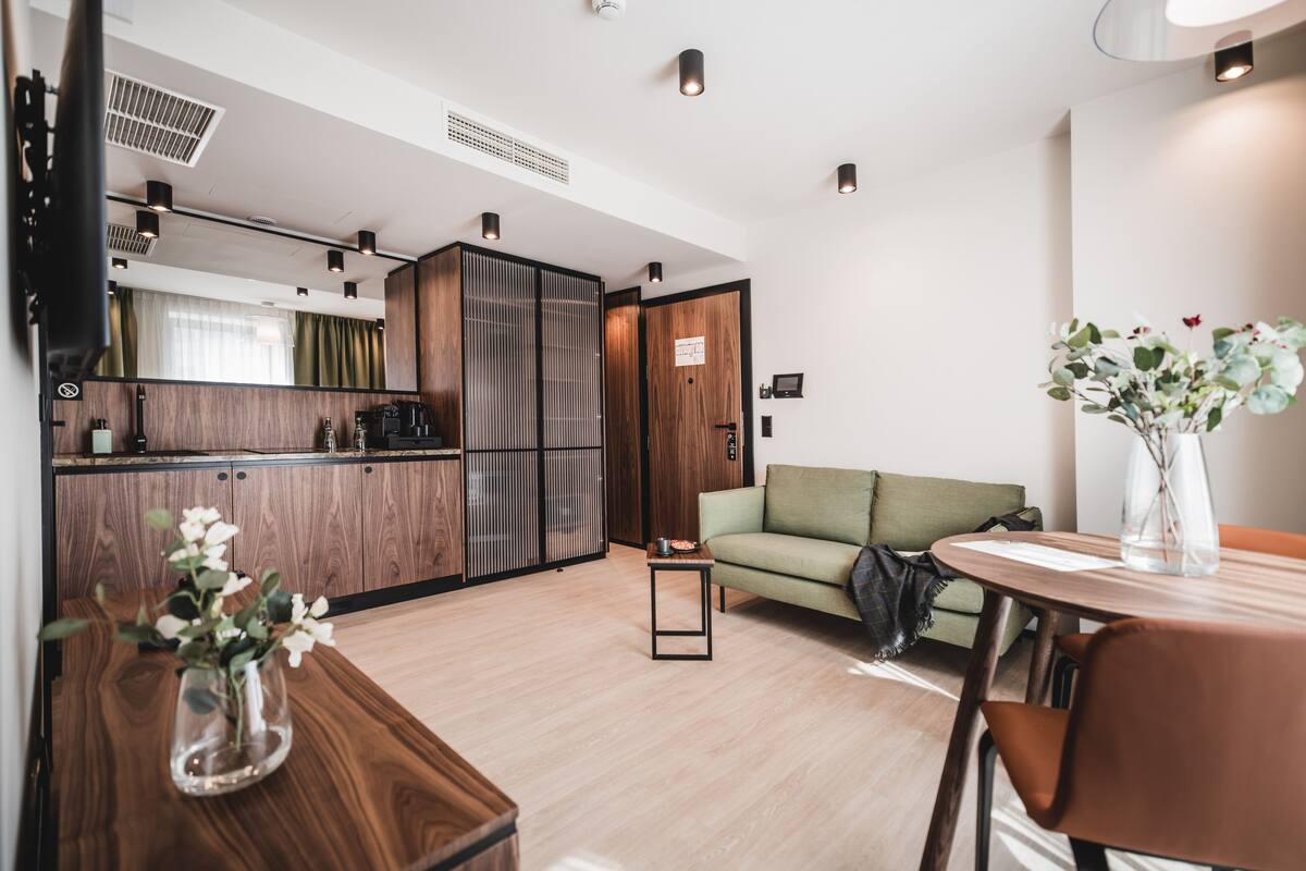 A modern kitchen with wooden cabinets and a built-in refrigerator. There is a dining area with a table and chairs, and a sofa in the living room.