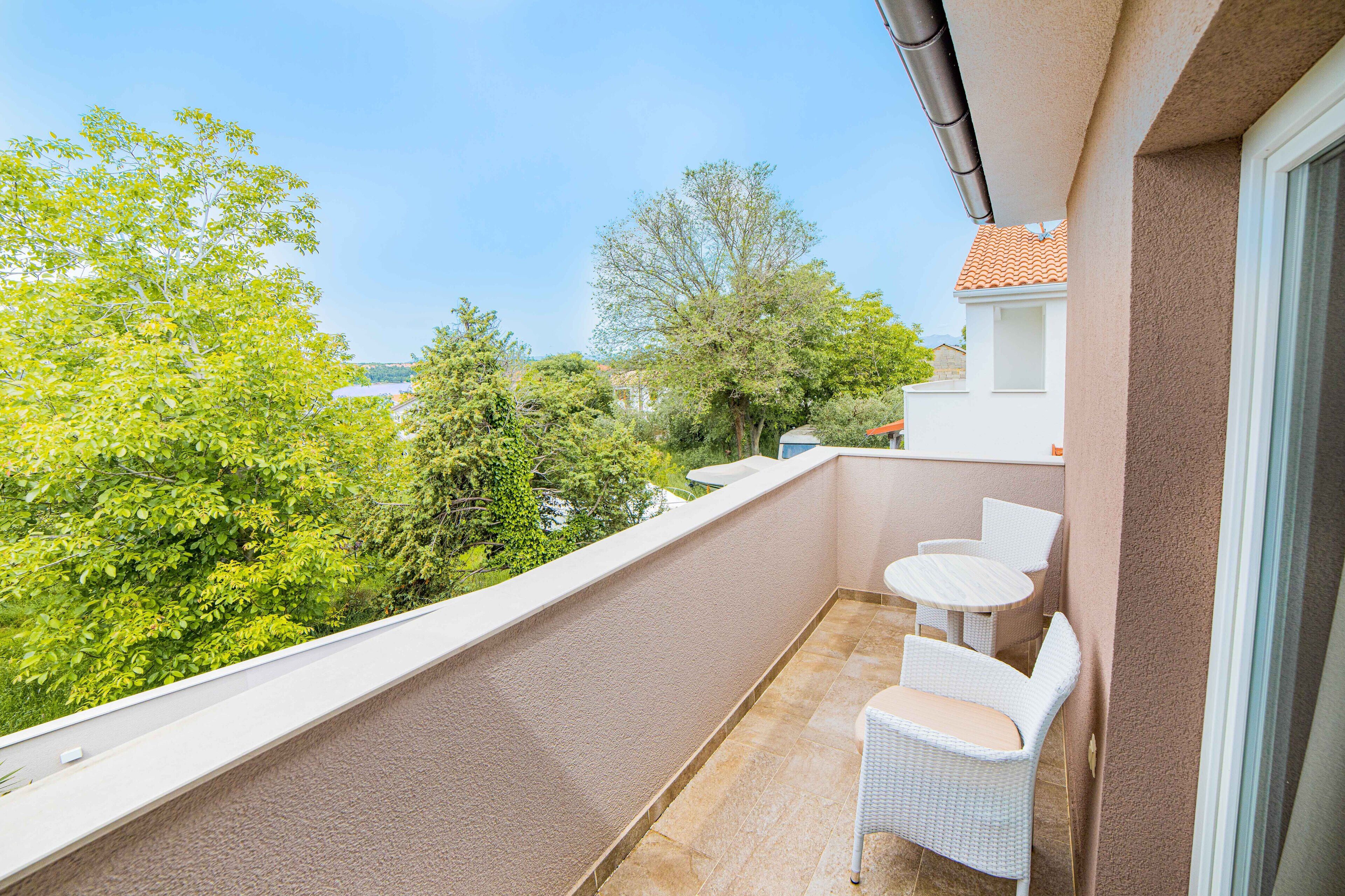 Un balcon doté de meubles en osier blanc, offrant une vue sur la verdure et les bâtiments, et dont le sol est recouvert de carreaux.
