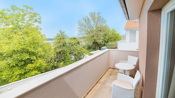 Un balcon doté de meubles en osier blanc, offrant une vue sur la verdure et les bâtiments, et dont le sol est recouvert de carreaux.