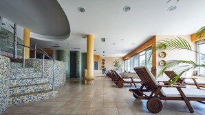 A spacious indoor area with tiled flooring, a staircase with mosaic tiles, wooden lounge chairs, and large windows allowing natural light. - Pohorje Village Wellbeing Resort – Forest Apartments Videc (Maribor)