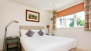 A neatly made bed in a room with a window, a framed picture on the wall, and a lamp on a nightstand.