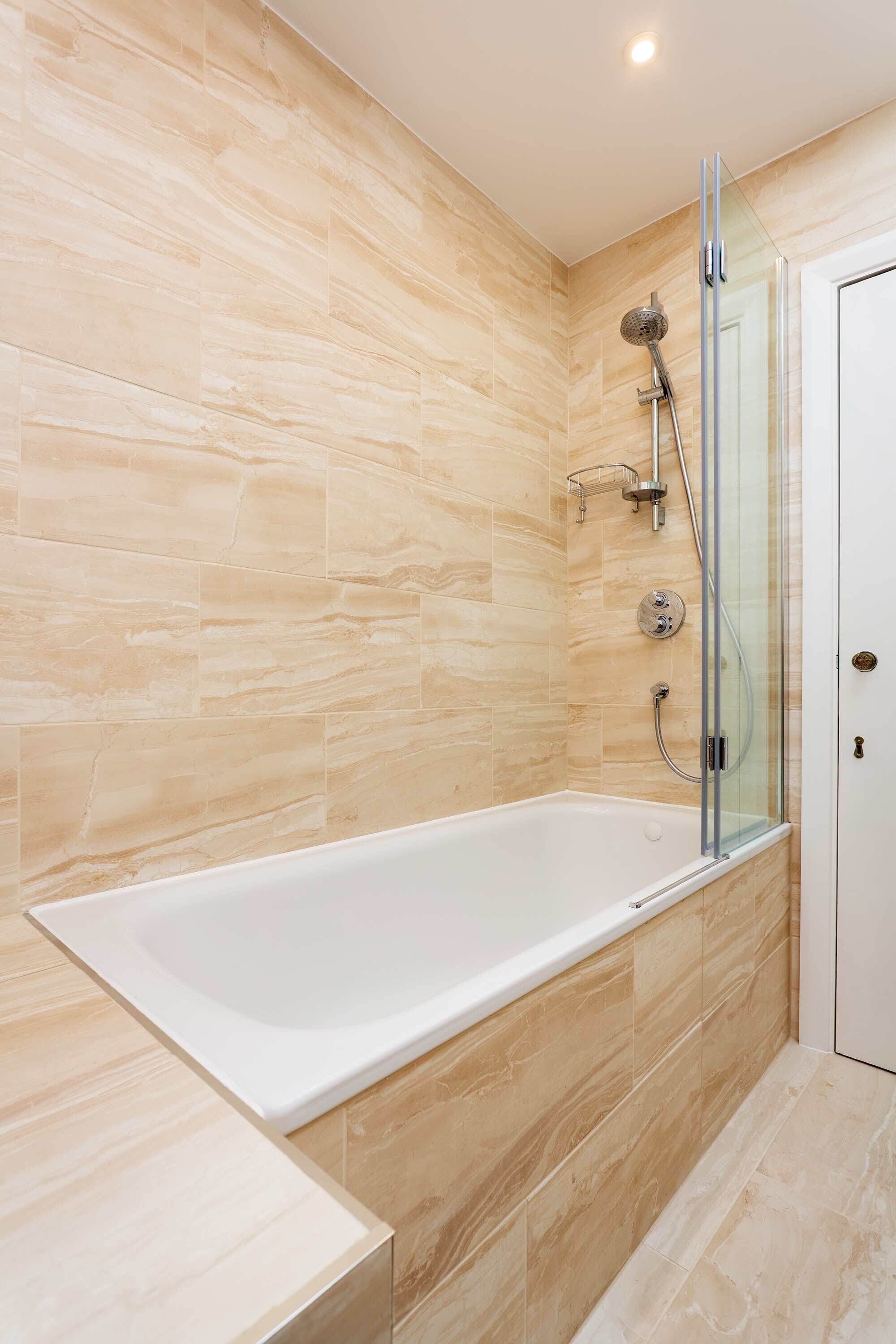 A modern bathroom with a glass-enclosed shower, a white bathtub, and beige marble tiles.