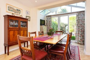 A dining area with a wooden table, red cushioned chairs, a glass cabinet, and a large window with curtains. - Clareville Classic, by Veeve (London)
