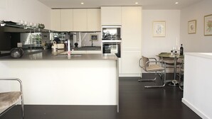 A modern kitchen with a central island, stainless steel appliances, and a dining area with a glass table and metal chairs. - Modern Edwardian, by Veeve (London)