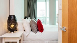 A hotel room with a bed, a bedside table with a lamp, and a view of a building through a window.